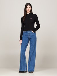 Donna in piedi che indossa un maglione aderente nero con zip e pantaloni jeans blu a vita alta e gamba larga con scarpe nere, sfondo neutro.