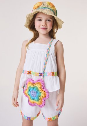 Jeune fille portant une tenue blanche sans manches décorée de motifs floraux colorés, un chapeau de soleil au crochet et un sac multicolore en forme de fleur.