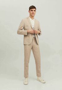 Noor mees beežis ülikonnas ja valges polo särgis, kinnitamas ülikonna nuppe, seismas heledal neutraalsel taustal valgete spordijalatsitega.