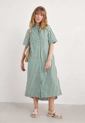 Gestreiftes grün-weißes Hemdkleid mit kurzen Ärmeln, asymmetrischem Saum und lockerer Passform. Getragen mit hellrosa Sandalen und einer geflochtenen Tasche.
