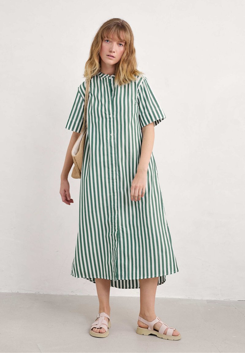 Abito a righe verde e bianco con bottoni, maniche corte, orlo asimmetrico e vestibilità comoda. Indossato con sandali rosa chiaro e una borsa intrecciata.
