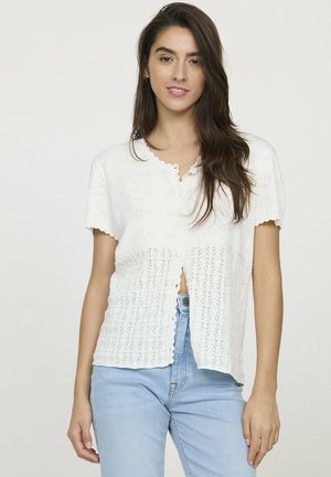 Jeune femme avec de longs cheveux bruns, portant un gilet en tricot blanc à manches courtes et un jean bleu clair, se tenant devant un fond uni.