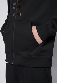 Felpa nera con zip, caratterizzata da una texture liscia, tasche laterali e accenti metallici in bronzo e grigio sul davanti per creare contrasto.