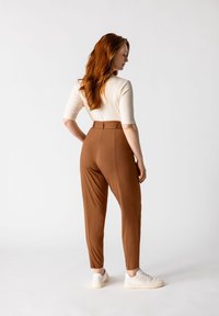 Pantalones marrones de talle alto con una textura suave y perneras estrechas, combinados con una blusa crema de manga tres cuartos y zapatillas blancas.