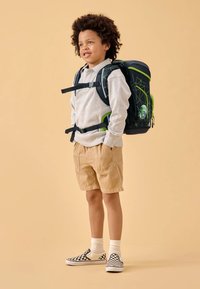 La mochila presenta un material oscuro con acentos verdes, patrones geométricos y un bolsillo lateral con un gráfico de un personaje. Se lleva con pantalones cortos beige.