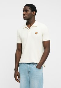 Hombre con camisa polo blanca de manga corta con un pequeño emblema de zorro, jeans azul claro, de pie con una mano en el bolsillo, mirando hacia la izquierda.