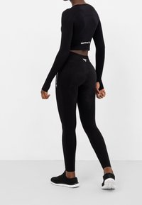 Černý sportovní set obsahující crop top s dlouhým rukávem a legíny s vysokým pasem, obojí vyrobené z texturované látky a doplněné logem. Černé tenisky celý look dokončují.