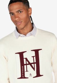 Suéter crema con diseño gráfico burdeos de "H" y "N". Presenta un cuello redondo y puños acanalados, superpuesto a un collar de camisa a rayas.