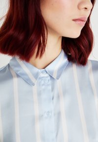 Chemise en soie bleu clair avec des rayures verticales blanches, dotée d'un col pointu et de deux petits boutons sur la patte de boutonnage. Texture lisse.