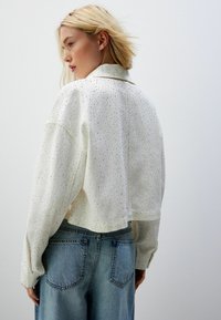 Afkortet hvid jakke med struktureret design og små metaludsmykninger. Har sænket skuldre og lange ærmer, parret med blå jeans.