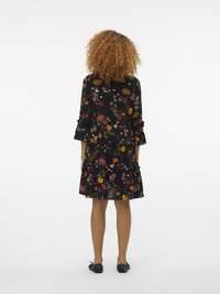 Vestido preto com flores, com mangas balonê e saia em camadas, apresentando padrões coloridos de flores em amarelo, vermelho e verde. Sapatos planos usados.