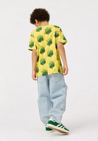 Żółta i zielona koszulka w sześciokątny wzór, luźny krój, dopasowana z jasnoniebieskimi, szerokimi dżinsami i zielonymi sneakersami z białymi paskami.