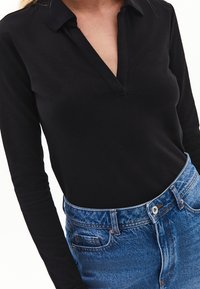 Schwarzes langärmliges Polohemd mit V-Ausschnitt, kombiniert mit hoch geschnittenen blauen Denim-Jeans, die über einen Knopf und Taschen verfügen.