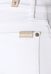 Los jeans de mezclilla blancos presentan una etiqueta de cuero marrón claro con "for all mankind" grabado, un botón de metal y costuras en contraste en la cinturilla.