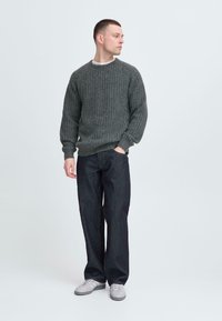 Grå ribbet strikkegenser med rund hals, parret med løse mørke denimjeans og lyse grå joggesko, som står mot en ensfarget bakgrunn.