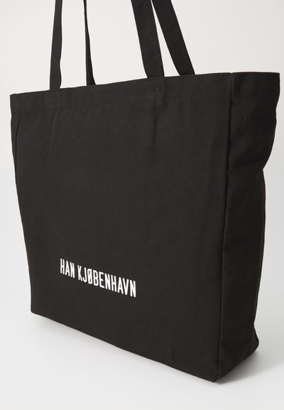 Geantă de umăr neagră din pânză, cu două mânere. Fața prezintă text alb care spune "HAN KJØBENHAVN." Formă rectangulară și textură moale.