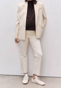 Blazer beige doppiopetto sopra una maglietta marrone, pantaloni bianchi cropped e sneakers bianche. Tessuto morbido, design minimalista, accenti sottili.