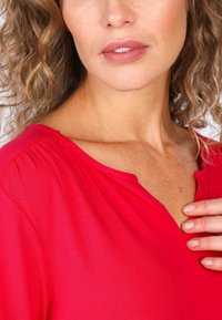 Rotes, kurzärmliges Oberteil aus weichem Stoff. Hat einen V-Ausschnitt und subtile Falten an den Schultern. Glatte Textur.