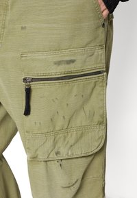 Close-up of green cargo pants pocket with black zipper and faint dark stains, hand partially in side pocket against white background.