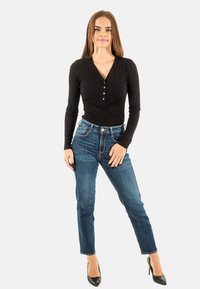 Haut noir à manches longues côtelé avec détail de boutons, associé à un jean en denim bleu et des talons hauts noirs ; le modèle se tient devant un fond neutre.