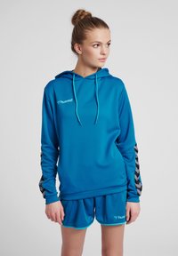 Blå sportig huvtröja med en strukturerad yta och kontrasterande svart chevronsmönster på ärmarna. Matchas med shorts som har logotyp.