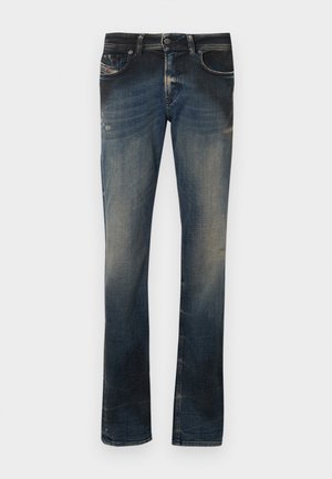 Jeans de denim azul escuro com um leve desbotado, apresentando cinco bolsos, fecho com zip e costuras em destaque. Corte reto com ligeiras desgastes.