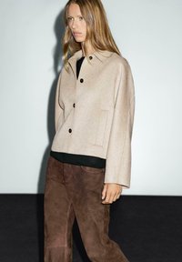 Beige wool blend jacket with a cropped design, black buttons, and a chest pocket, paired with brown suede pants. Smooth texture overall.