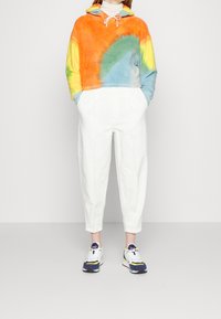 Felpa corta tie-dye in arancione, giallo, verde e blu, abbinata a pantaloni bianchi affusolati. Sneakers bianche con dettagli multicolore.