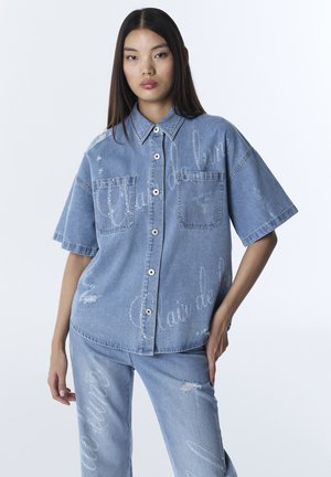 Frau in locker sitzendem hellblauem Jeanshemd und Hose mit ausgefransten Details und weißem Schriftzug, die vor einem schlichten weißen Hintergrund posiert.