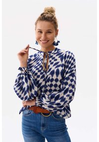 Witte blouse met blauwe onregelmatige patronen, geraffineerde ballonslaven en een strikhalslijn. Gecombineerd met hoge taille blauwe jeans en een bruine riem.