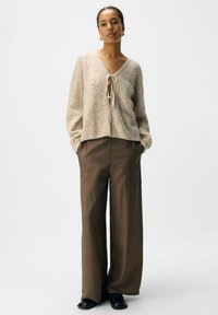 Vrouw die staat met handen in de zakken, gekleed in een beige gebreide cardigan die aan de voorkant is gestrikt, bruine wijde broek en zwarte schoenen, tegen een witte achtergrond.