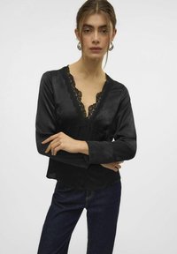 Sort bluse med lange ærmer, dyb V-udskæring dekoreret med blonde. Blød, glat tekstur, lagdelt design, parret med mørkeblå denim.