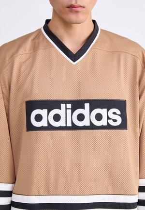 Osoba nosząca beżową sportową koszulkę Adidas z czarno-białym obszyciem i dużym logo Adidas na piersi.