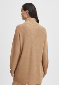 Pull en maille marron clair avec un col montant, présentant un motif côtelé texturé et des détails diagonaux au dos. Coupe oversize.