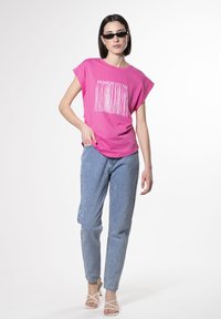 Lyserød t-shirt med korte ærmer med teksten "FASHION VICTIM" og frynsedesign, parret med lyseblå, spraglet denimjeans og hvide strophæle.