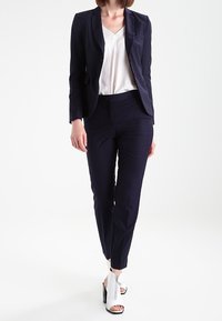 Mörkblått figursytt kavaj med en enda knapp, matchas med avsmalnande byxor och en vit v-ringad topp. Skorna är högklackade med dragkedja.