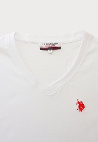Tricou alb din bumbac cu decolteu în V, având un logo roșu brodat pe partea stângă. Eticheta spune "U.S. Polo Assn." cu mărimea "M." Textură netedă.