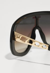 Occhiali da sole neri oversize con lenti sfumate, accentuati da dettagli dorati sulle tempie con ritagli circolari e un design elegante.