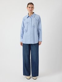 Light blue striped button-up shirt featuring floral embroidery, worn with wide-leg dark denim jeans and white heeled shoes. Plain background.