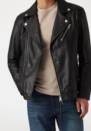 Uomo che indossa una giacca da motociclista in pelle nera con dettagli di cerniera e bottoni argentati sopra un maglione beige lavorato e jeans blu.