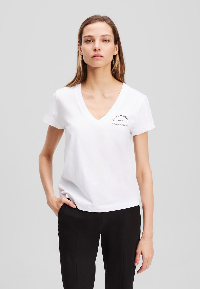 Weißes Baumwoll-T-Shirt mit V-Ausschnitt, kurzen Ärmeln und einem erhabenen schwarzen Logo auf der Brust, kombiniert mit schwarzen Hosen.