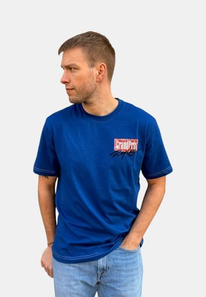 Joven con pelo corto que lleva una camiseta azul "Grand Prix Replay" y vaqueros azul claro, mirando hacia su izquierda con una mano en el bolsillo.