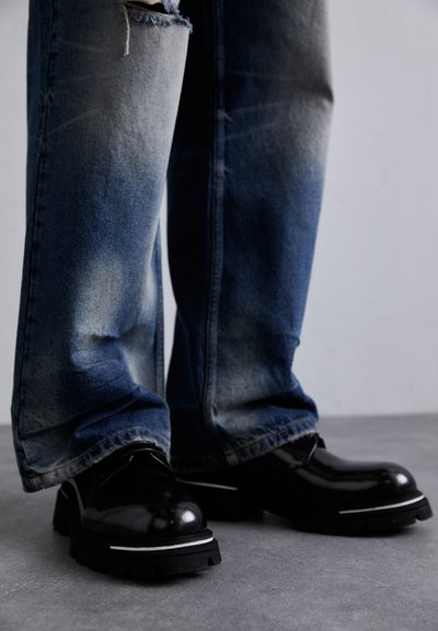 Schwarze Lederschuhe mit glänzender Oberfläche, featuring eine dicke Sohle und einen weißen Streifen. Getragen mit abgewetzten blauen Jeanshosen, die leichte Verblassungen zeigen.