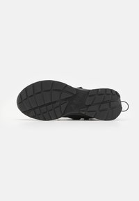 Semelle de sneaker noire avec une surface texturée et patternée, dotée de rainures profondes pour une meilleure adhérence et d'un logo en relief au niveau du milieu.