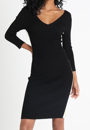 Sort ribstrikket kjole med lange ærmer og V-hals. Figursyet silhuet med sideslids og kontrasterende lysegule detaljer.
