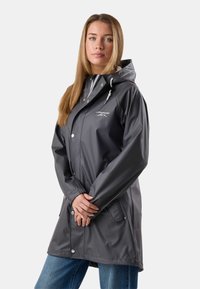 Vrouw met lang haar, gekleed in een donkergrijze waterdichte jas met capuchon en witte trekkoorden, staand met de handen gevouwen voor zich.