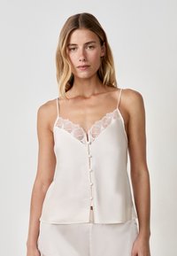 Camisola di seta color crema con bordo in pizzo lungo il colletto, presenta una chiusura a bottoni sul davanti e spalline sottili, offrendo una silhouette morbida e fluida.
