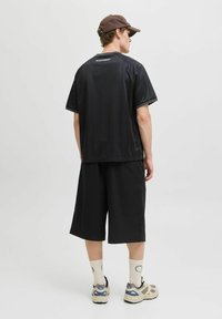 Chemise noire à manches courtes, coupe ample, et shorts assortis jusqu'aux genoux, fabriqués en tissu lisse. Portés avec des baskets beiges et grises et des chaussettes blanches.