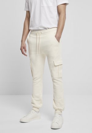 Kremfargede cargo joggebukser laget av myk stoff, med lommer på siden, elastiske mansjetter og snor i livet. Brukt med hvite sneakers.