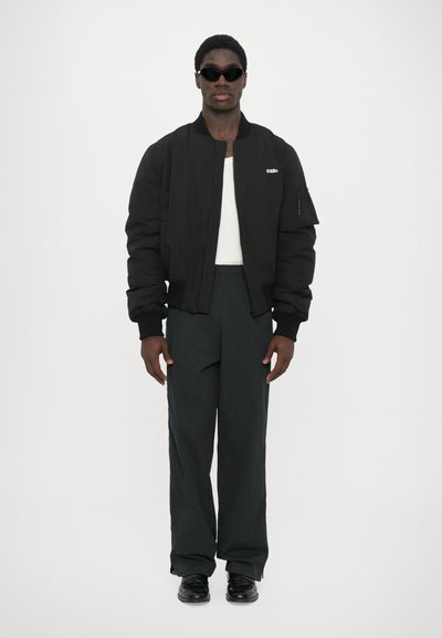 Schwarze Bomberjacke mit gerippten Bündchen, weißem Logo, enganliegenden schwarzen Hosen und schwarzen Schuhen, getragen mit schwarzen Sonnenbrillen und einem weißen Oberteil.